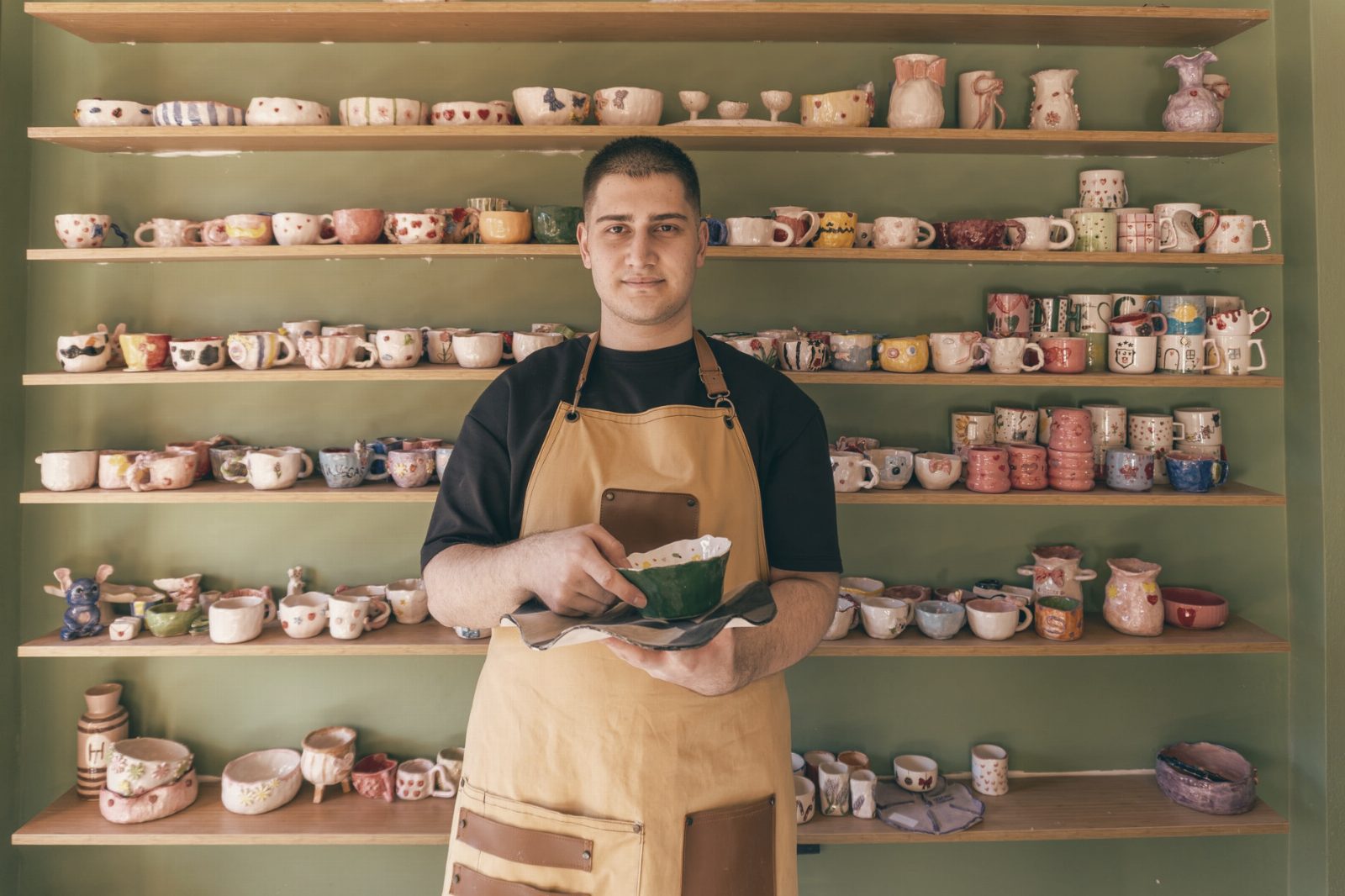 A ceramic artist presents a handcrafted mug in front of a shelf filled with colorful pottery.
