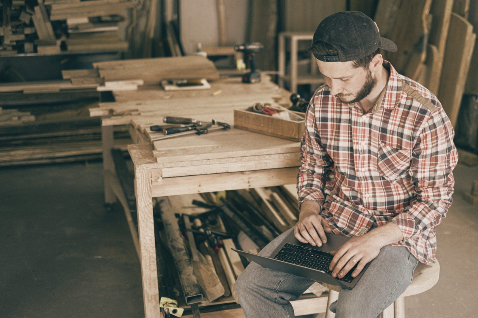 A carpenter working on his laptop in a wood workshop, surrounded by tools and materials.