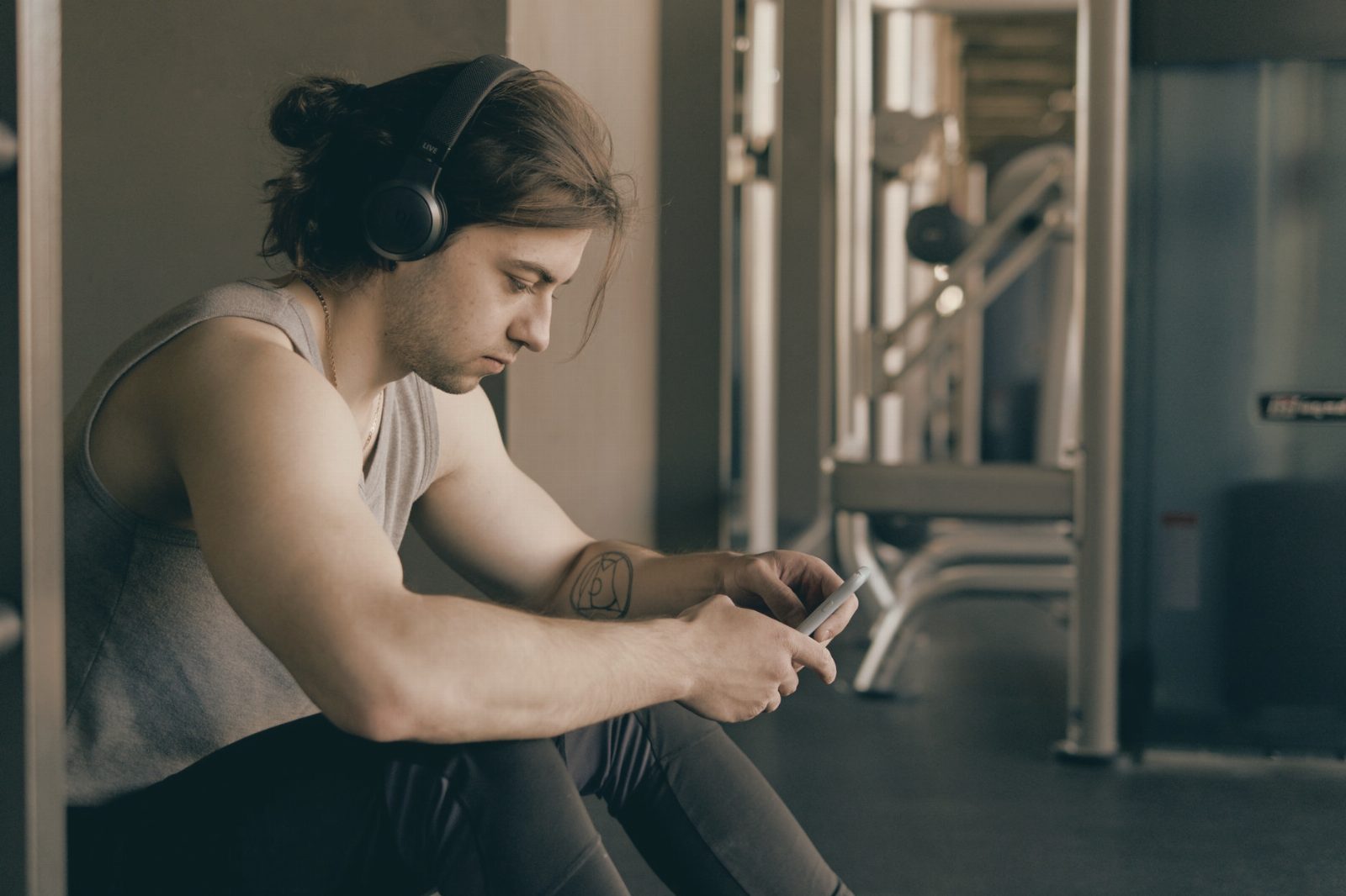 A young man wearing headphones sits in a gym, focused on texting with his smartphone.