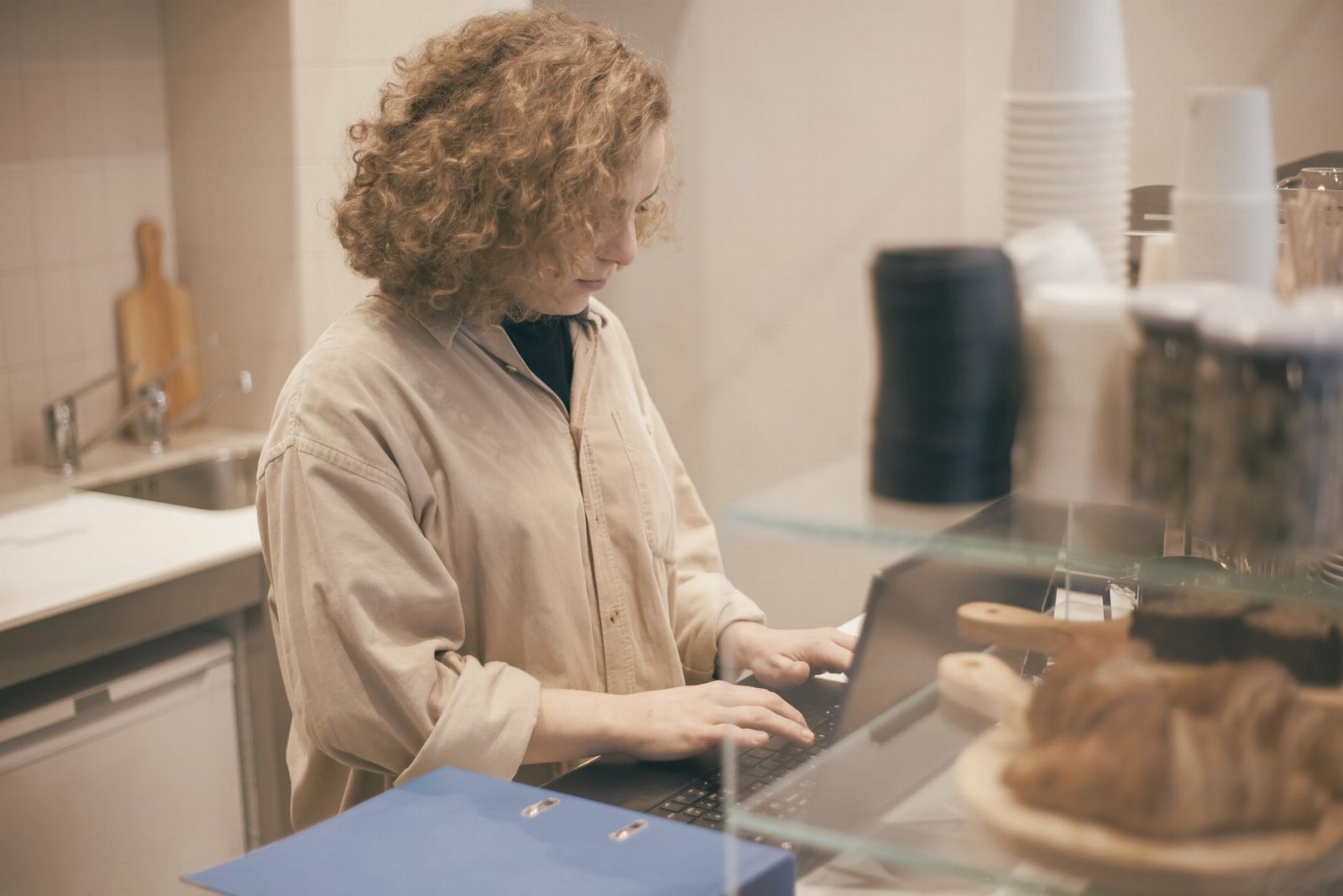 A young woman uses a laptop behind a café counter with pastries in foreground.