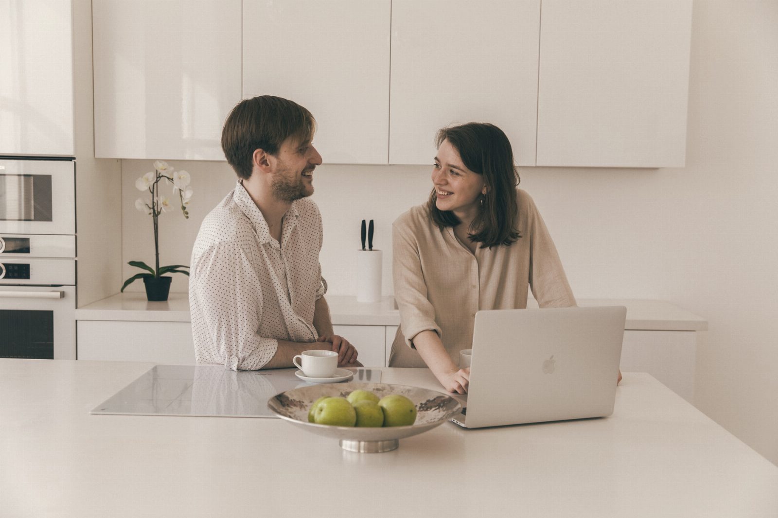 A smiling couple enjoying coffee together in a bright kitchen setting with a laptop and fruit.