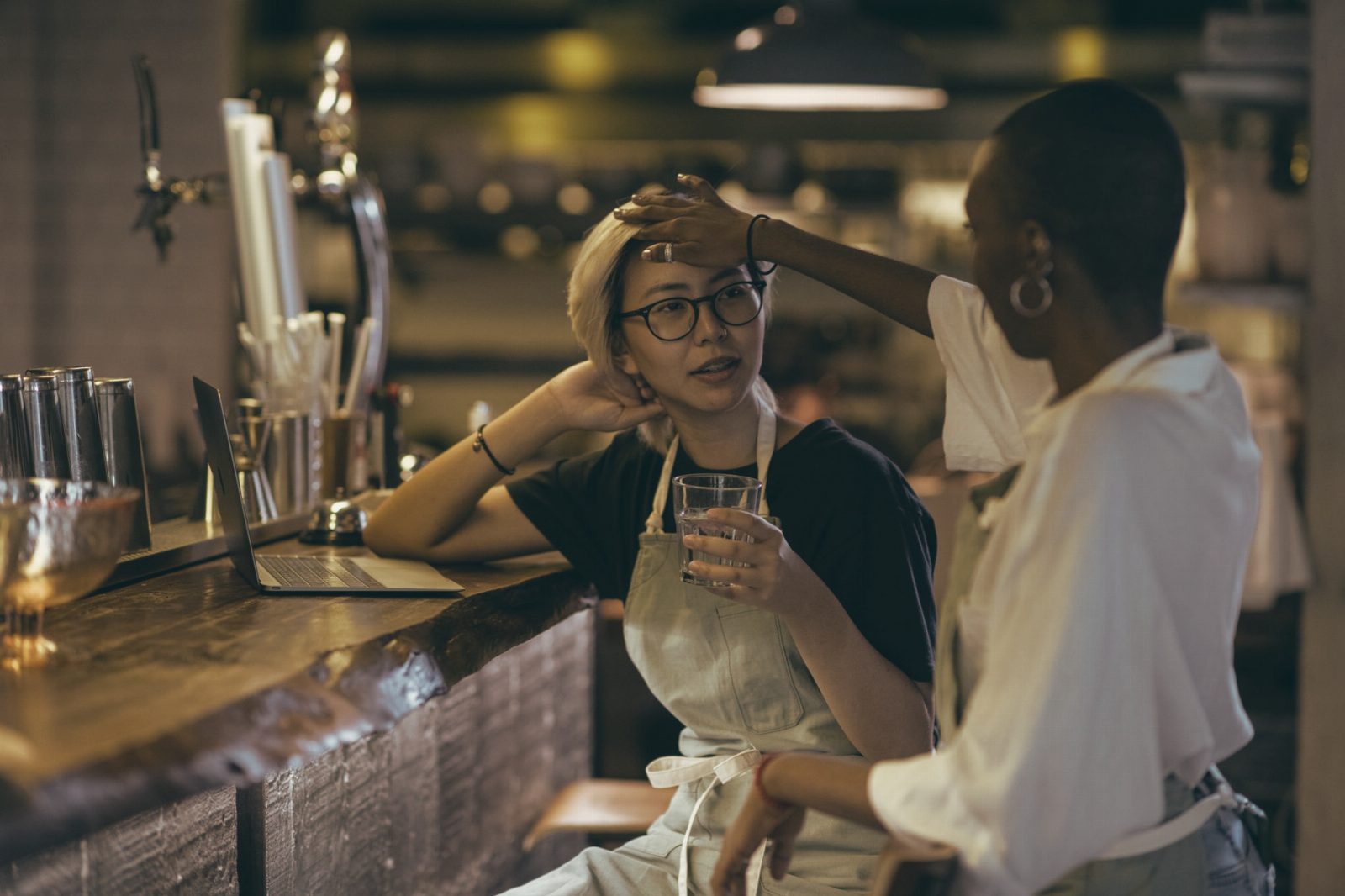 Positive diverse waitresses in aprons gathering at modern bar counter with drink and netbook while chatting and touching hair