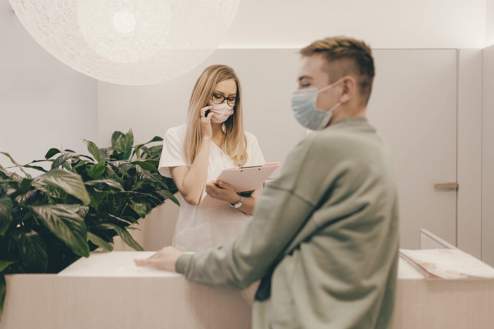 Healthcare worker and patient wearing masks at clinic front desk during pandemic, highlighting safety measures.